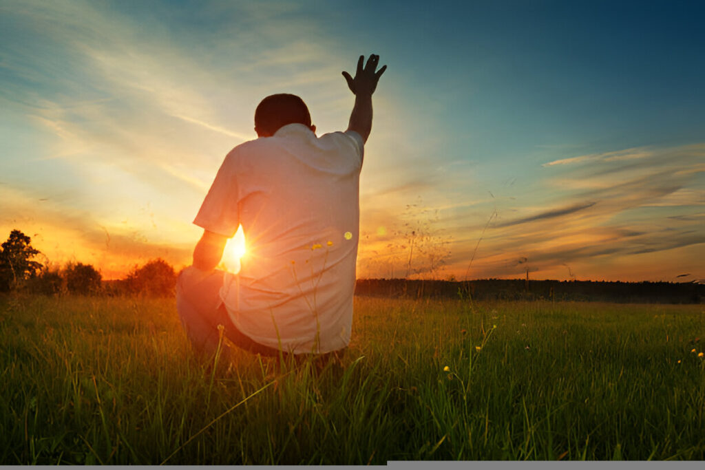 Man kneeling in a grassy field at sunset with hand lifted in worship to God.