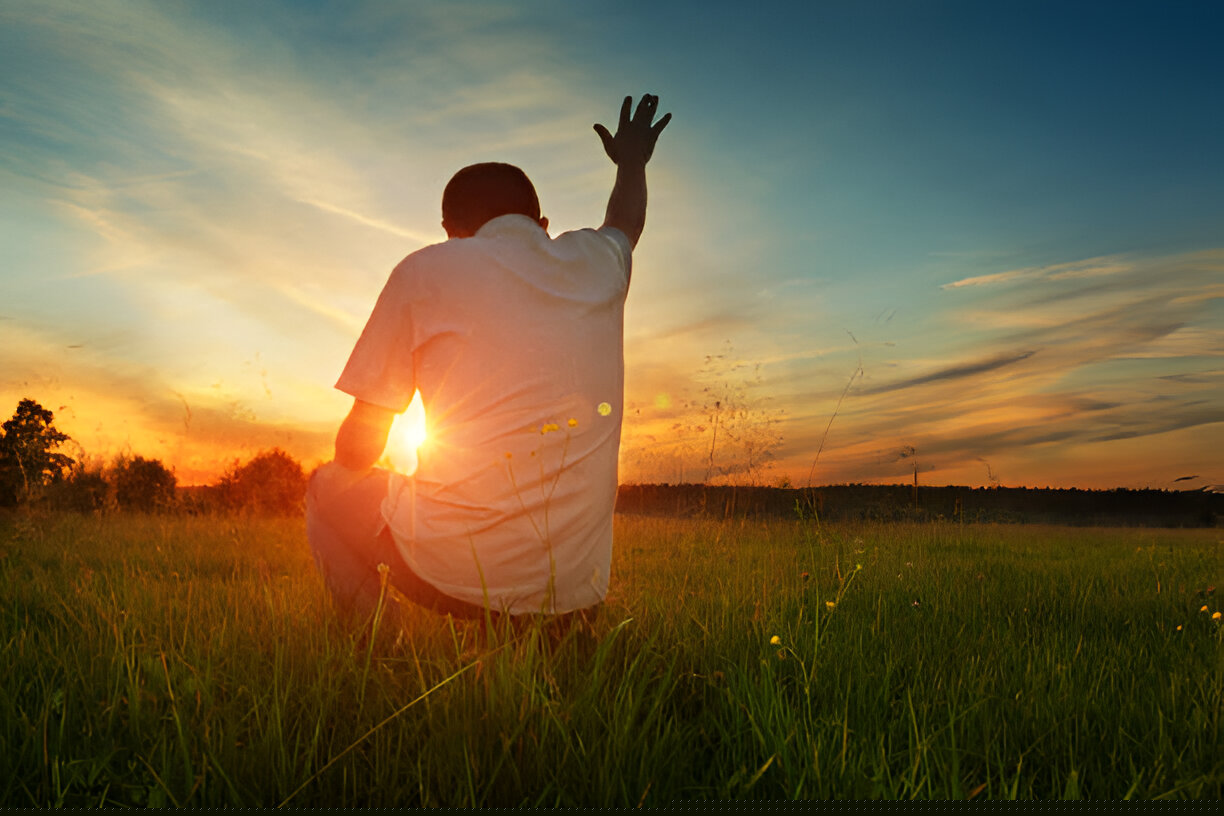 Man kneeling in a grassy field at sunset with hand lifted in worship to God.